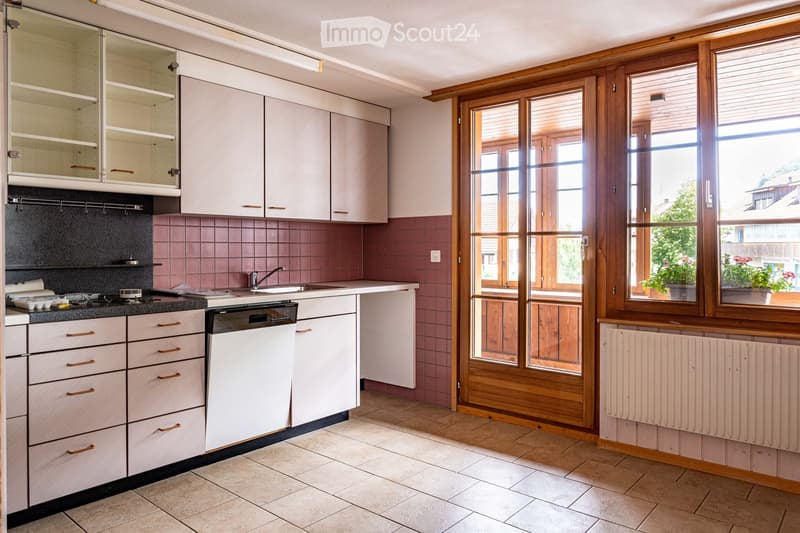Bright kitchen with white cabinets, pink tiled backsplash, and wooden window frames. Features a sink, dishwasher, and radiator.