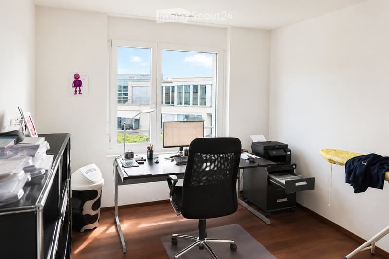 A modern, well-lit office space with a wooden floor, large windows, a desk with a computer setup, a comfortable office chair, and a printer. The room also includes a trash can, a cabinet, and a lamp.