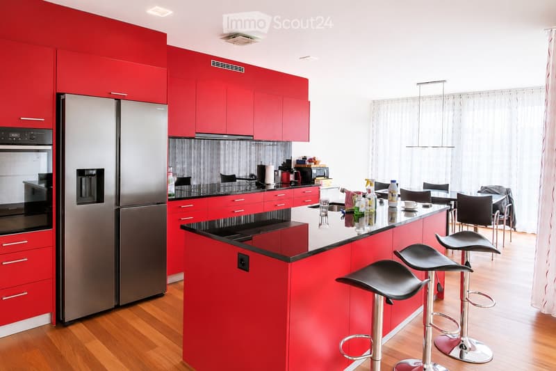 Modern kitchen with red cabinets, stainless steel appliances, island with bar stools, and dining area with chairs and table.