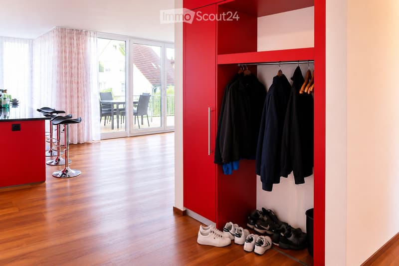 Modern hallway with wooden flooring, red wardrobe, several hanging coats, shoes on the floor, and a small seating area with stools and a table leading to a balcony.
