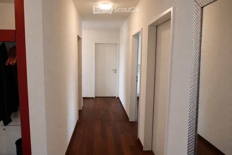 A narrow hallway with wooden flooring, white walls, and several doors leading to different rooms. A coat rack with hanging coats is visible on the left side, and a large mirror is on the right wall.