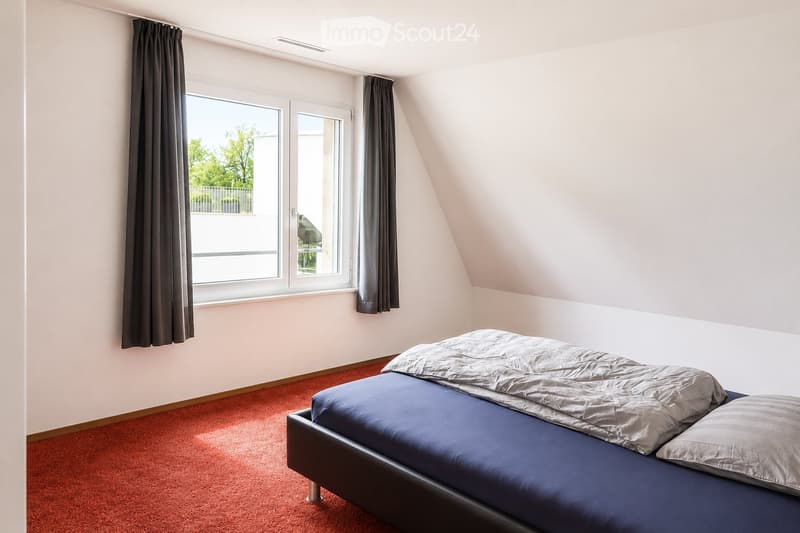 Bedroom with a single bed, blue bedding, and red carpet. Large window with dark curtains and a view of greenery outside.