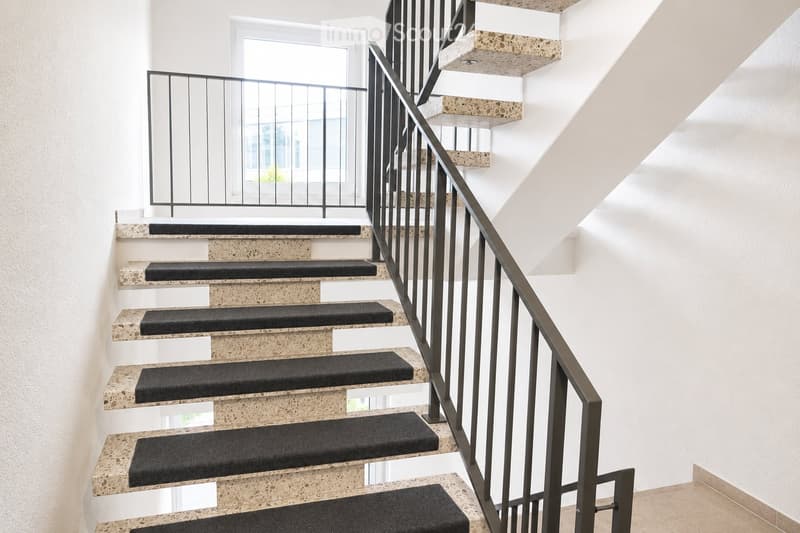 Modern staircase with beige stone steps and black carpet runners, featuring a sleek metal railing and a window at the top landing.