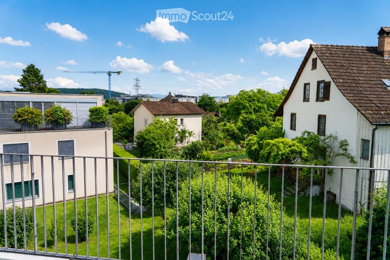 View from a balcony showing a well-maintained garden with green grass, bushes, and trees, surrounded by residential houses and a clear blue sky.