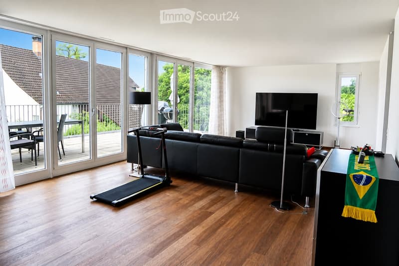Modern living room with large sliding glass doors leading to a balcony, a black sectional sofa, a treadmill, a TV, and a dining area with chairs and a table.