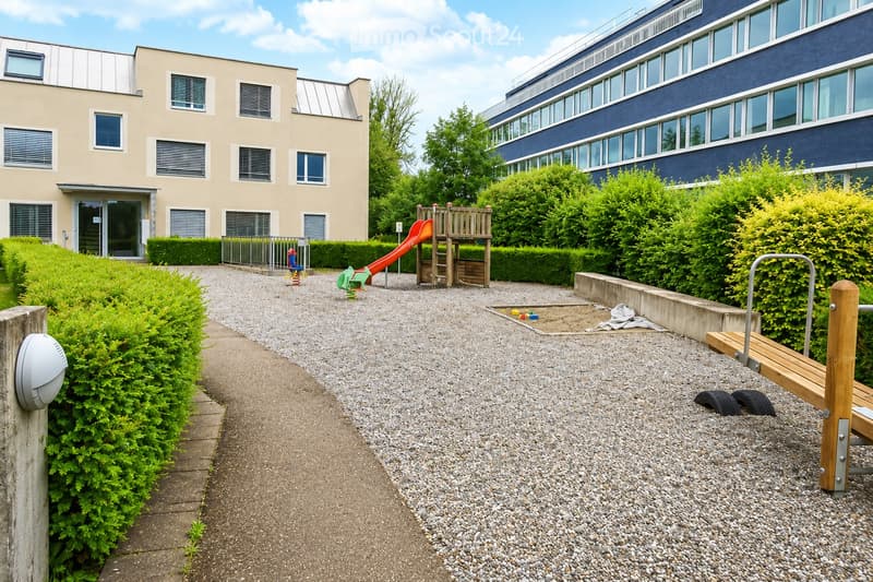 Outdoor playground area with a slide, climbing frame, and sandpit, surrounded by greenery and buildings.