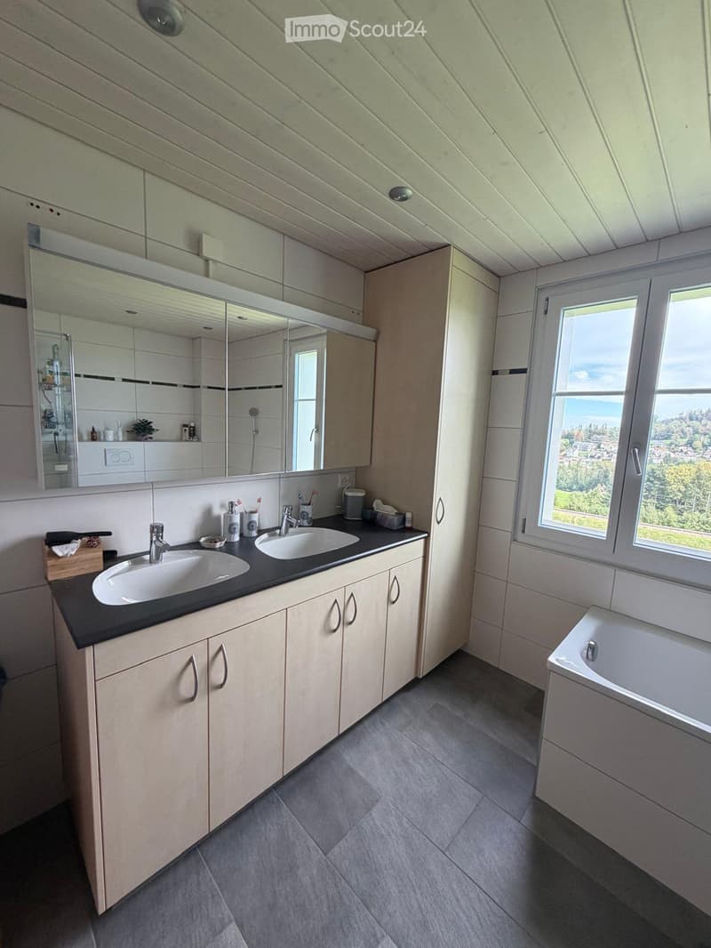 Modern bathroom with double sink, large mirror, bathtub, and ample natural light from a window overlooking a scenic view.