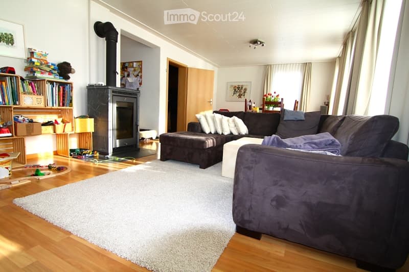 Living room with a dark sectional sofa, coffee table, fireplace, bookshelf, and dining area visible in the background.