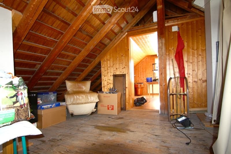 Attic space with wooden beams and exposed ceiling, featuring a cream-colored sofa, a small table, a chair, and several storage boxes.