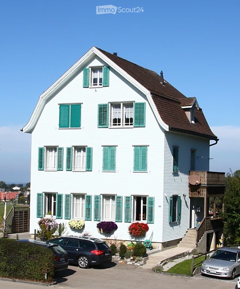 Two-story white house with green shutters, multiple windows, and a brown roof. There are flower boxes on the walls and a small garden area with plants. Three cars are parked in front of the house.
