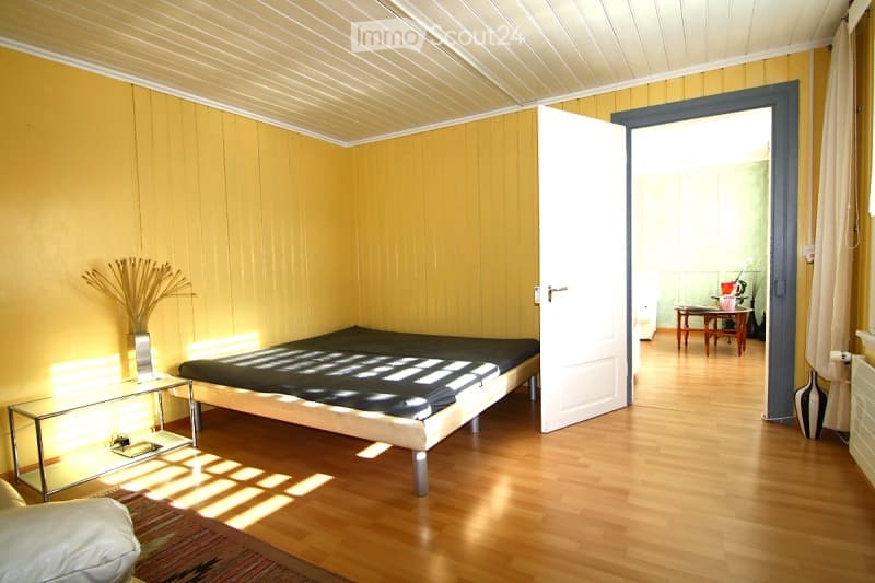 Bright bedroom with wooden walls and ceiling, featuring a bed, nightstand, vase with dried plants, and an open door leading to another room with a small table and chair.