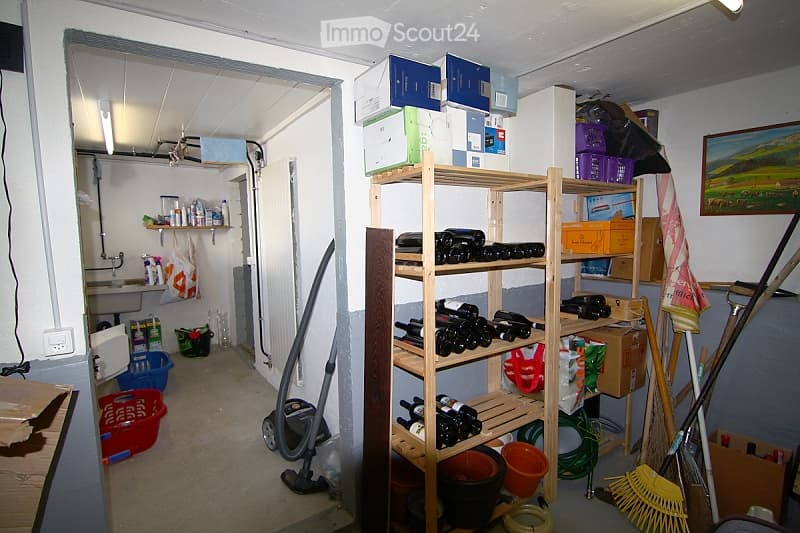 A cluttered storage room with shelves holding wine bottles, cleaning supplies, and miscellaneous items. There are also buckets, baskets, and a vacuum cleaner visible.