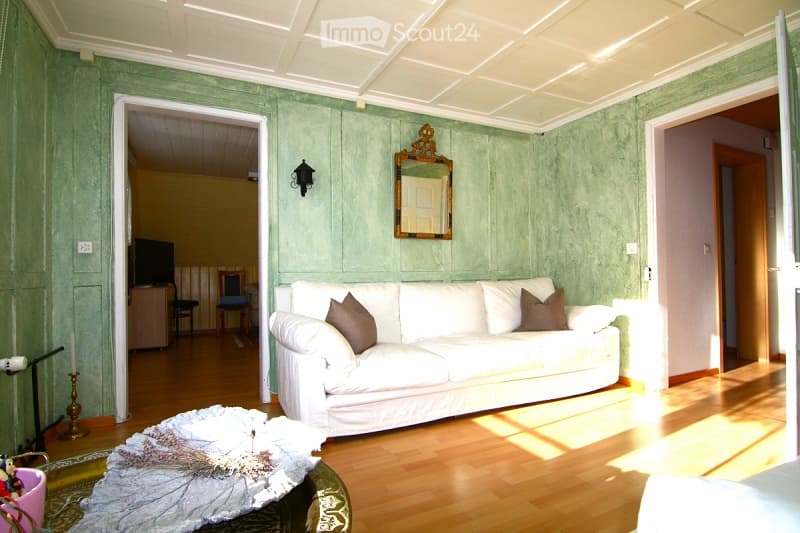 Living room with green textured walls, white ceiling, white sofa with throw pillows, coffee table with decorative items, mirror on wall, wall sconce, and visible adjacent room with TV and chair.