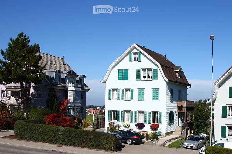 A two-story white house with green shutters and a brown roof, surrounded by greenery and parked cars.