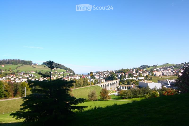 A panoramic view of a town with a mix of residential and commercial buildings, surrounded by green hills and a clear blue sky. A prominent aqueduct structure is visible in the distance.