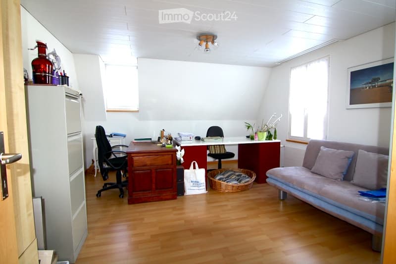 A multi-functional room with a wooden floor, a sofa, a desk with chairs, a refrigerator, and a wicker basket. The room is well-lit with natural light from large windows and has a cozy, lived-in feel.