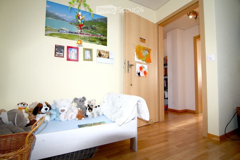 A cozy children's room with a small bed covered in stuffed animals, a wicker basket, and various picture frames and wall art on the walls. The room has wooden flooring and an open doorway leading to another area.