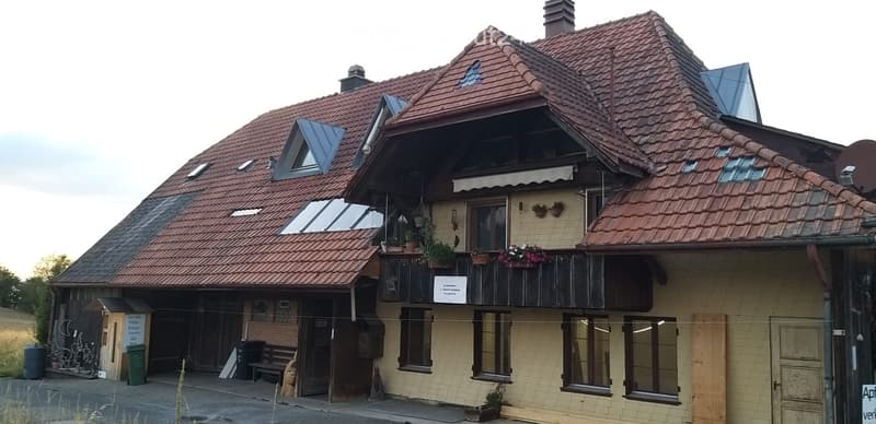 Two-story building with red-tiled roof, multiple windows, and a balcony adorned with potted plants. The facade features a mix of brick and plaster finishes.