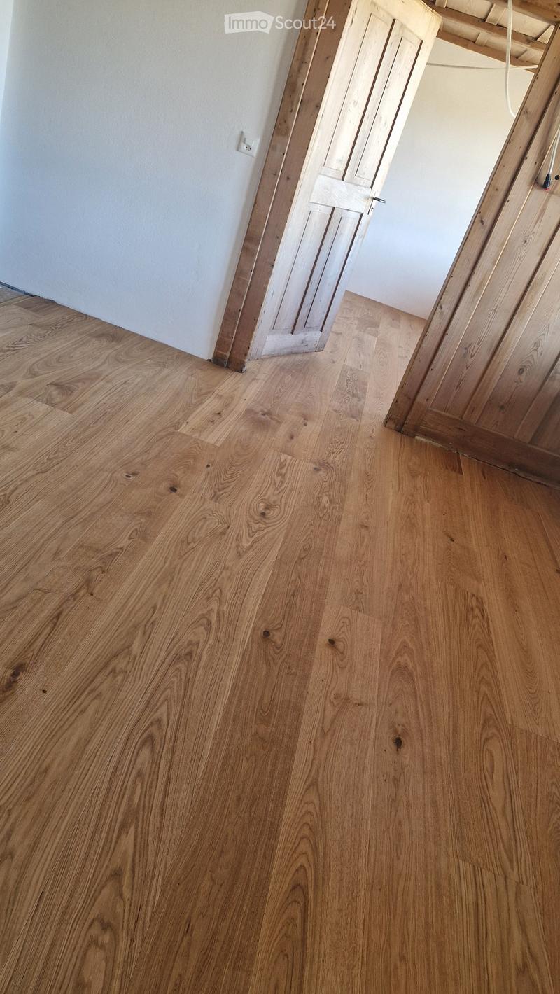 Empty room with light wooden flooring and white walls, featuring a partially open wooden door leading to another area.