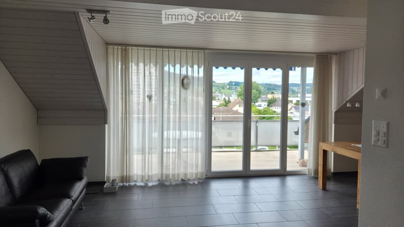 Modern living room with large sliding glass doors, sheer curtains, dark tiled floor, black leather sofa, and wooden table.
