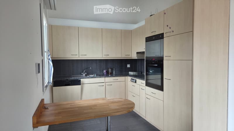 Modern kitchen with light wood cabinets, stainless steel appliances, and a wooden table.