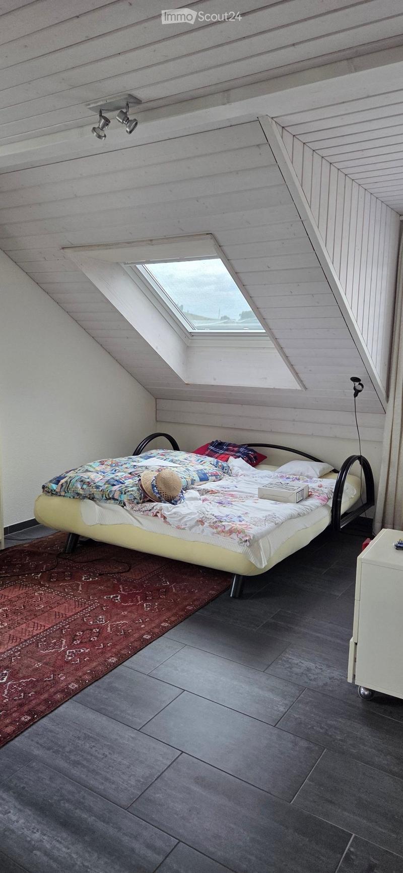 A modern attic bedroom with a slanted wooden ceiling, skylight window, grey tiled floor, and a colorful bed with a patterned duvet and pillows.