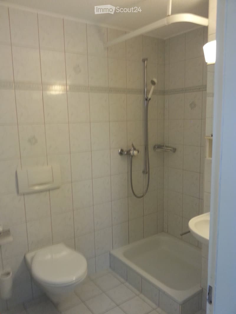 A small bathroom with a toilet, bathtub, sink, and a shower head with a handheld sprayer. The walls are tiled with light-colored tiles and a decorative border near the bathtub.