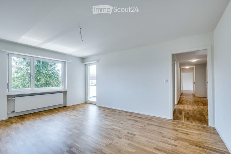 Empty room with light-colored wooden flooring, white walls, and large windows allowing natural light. The room features a radiator under one window and an open doorway leading to a hallway.