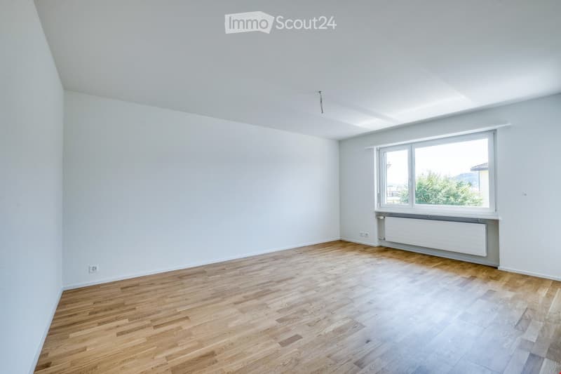 Empty room with light wooden flooring, white walls, and a large window with a view of greenery outside.
