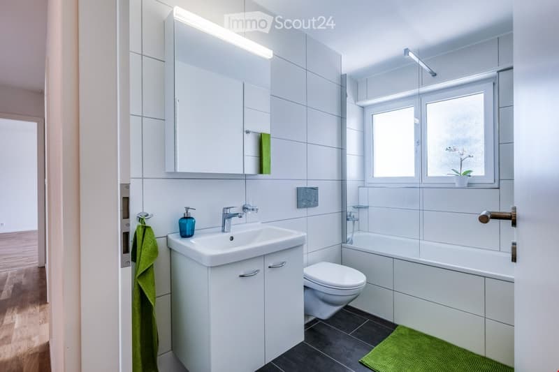 Modern bathroom with white tiles, a sink with a chrome faucet, a toilet, a bathtub, a large mirror cabinet, and a green towel on the side.