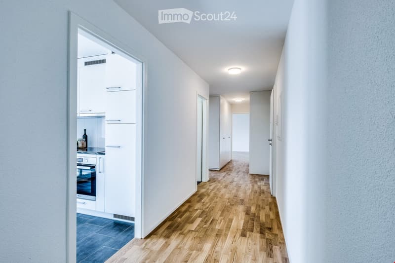 A modern hallway with wooden flooring, white walls, and recessed lighting. A kitchen area is visible to the left, featuring white cabinets, a refrigerator, and a stove.