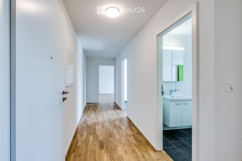 A clean, modern hallway with light wooden flooring, white walls, and a ceiling light. A partially visible bathroom with a sink, mirror, and light blue cabinet is seen to the right.