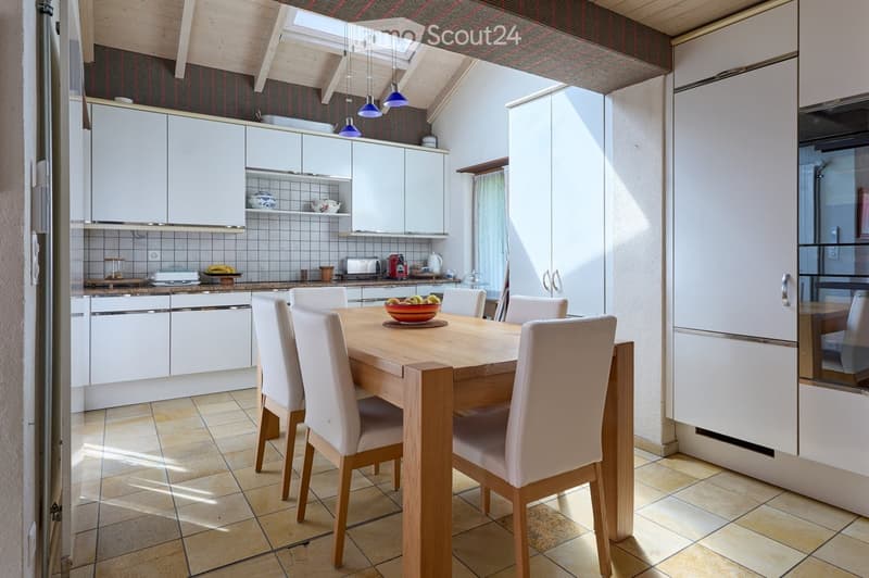 Bright kitchen with white cabinets, a wooden dining table with six chairs, and various appliances including a refrigerator, oven, toaster, and coffee maker. The room features a skylight and pendant lights.