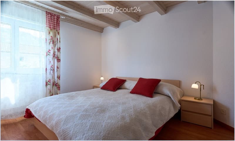 Cozy bedroom with wooden floor, white walls, and exposed wooden ceiling beams. Features a double bed with white bedding and red pillows, two nightstands with lamps, and floral curtains on the window.