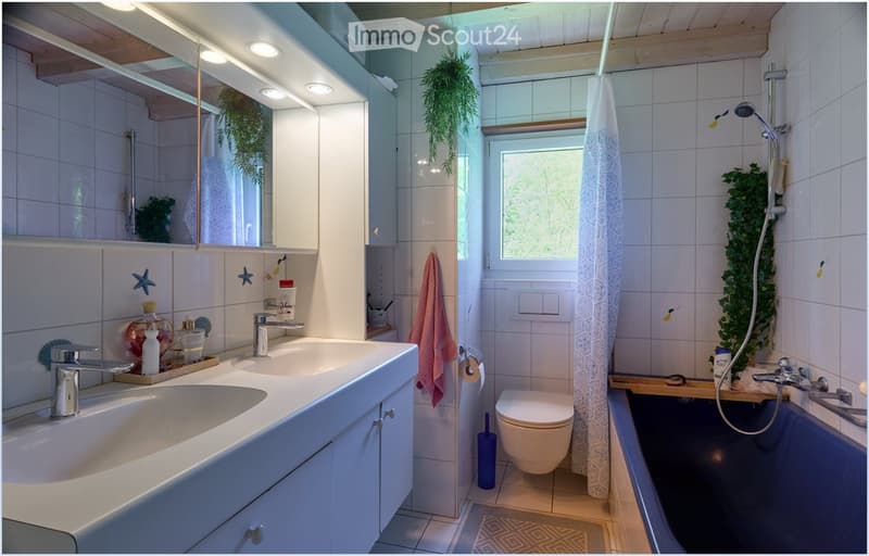 Modern bathroom with white tiles, double sink, bathtub, toilet, and a shower area with a curtain. Features a large mirror, decorative plants, and a towel hanging on the wall.