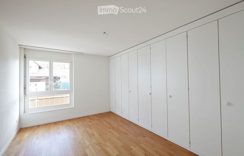 Empty room with light wooden flooring, white walls, large built-in white wardrobes, and a window with a view of a neighboring building and greenery.
