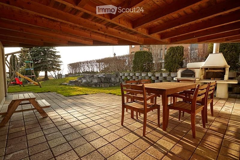 Outdoor patio with wooden table and chairs, stone flooring, wooden ceiling, and a view of a grassy yard with trees and a swing set.