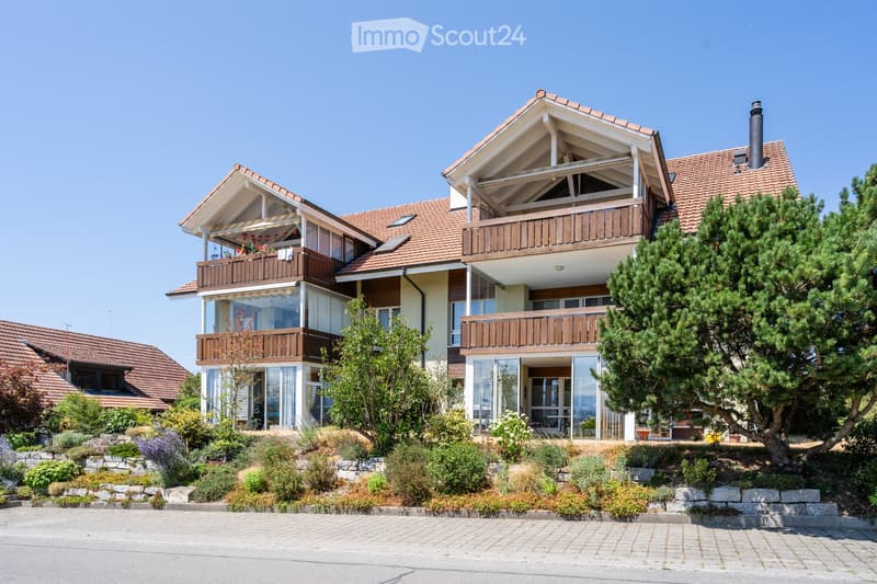 Two-story house with red-tiled roof, wooden balconies, large windows, and landscaped garden with trees and shrubs.