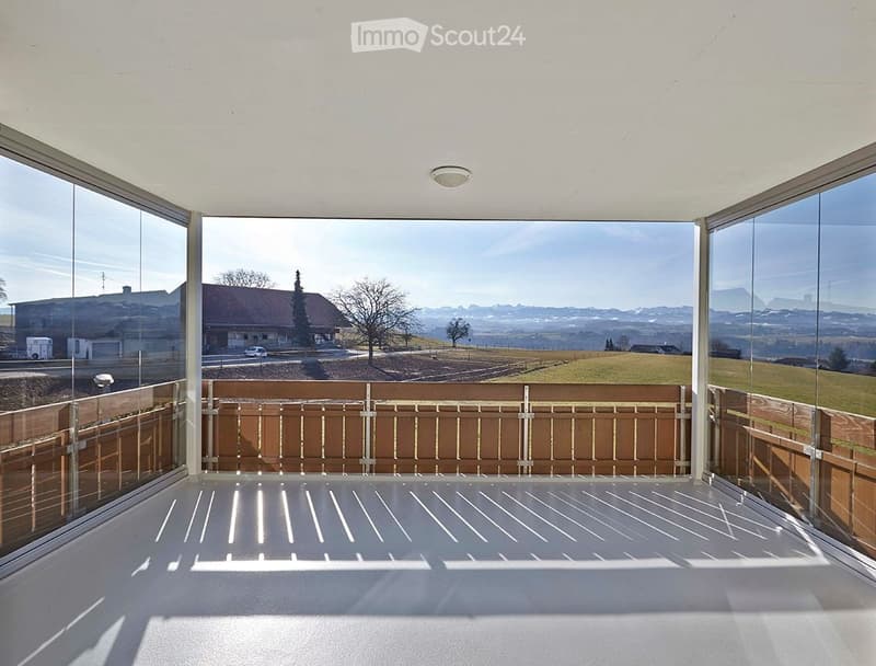 Modern balcony with large glass walls, wooden railing, and expansive scenic view of countryside and mountains.
