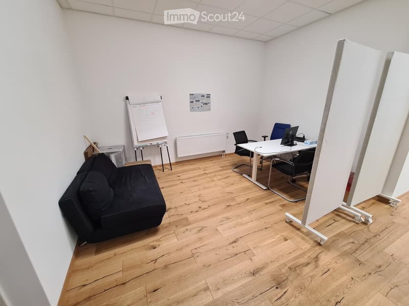 A modern, well-lit room with wooden flooring, a black sofa, a white table with chairs, a large screen, and a whiteboard on wheels.