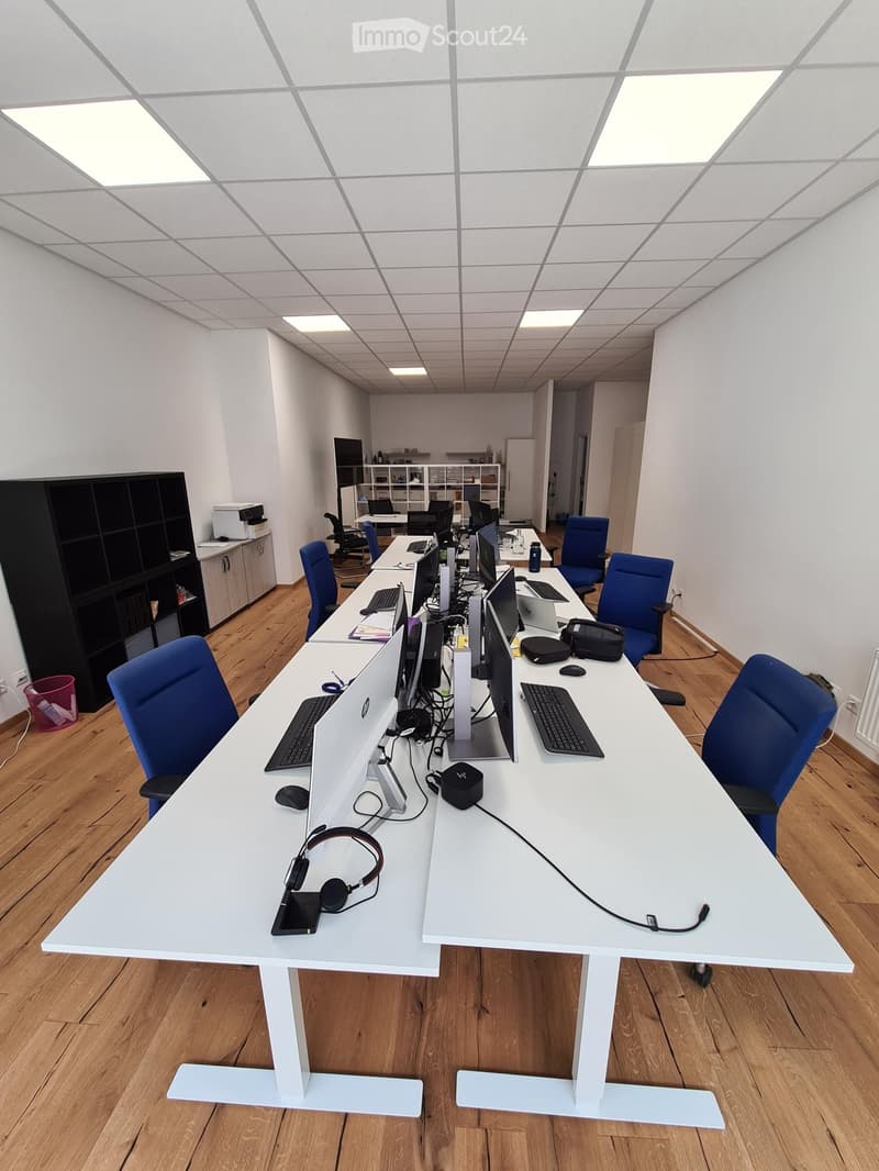 Modern office space with white desks, blue chairs, multiple computer setups, and various office equipment.