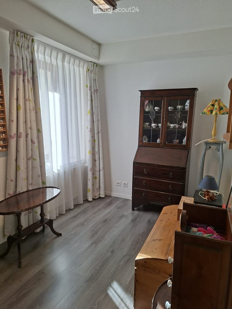 A well-lit room with wooden flooring, a round wooden table, a wooden cabinet with glass doors, a side table with a lamp, and floral curtains on the window.