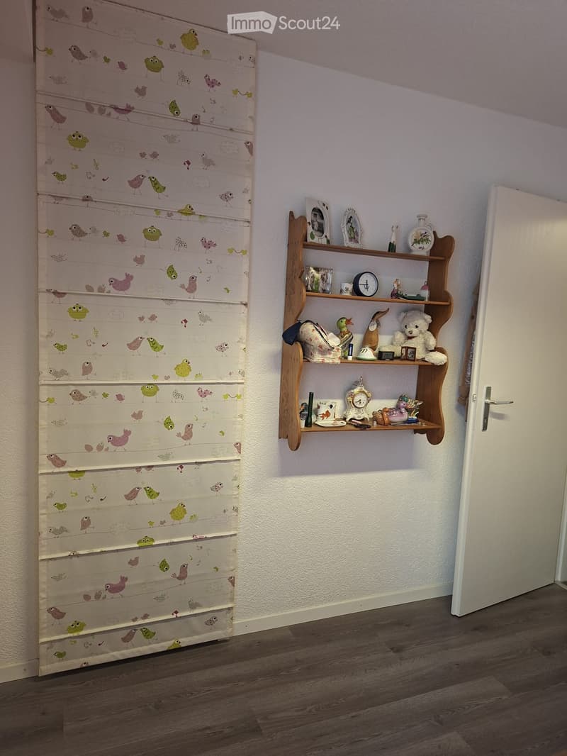A small, neatly arranged room with wooden flooring, a white door, and a wooden shelf holding various decorative items including a clock, a teddy bear, picture frames, and small toys.