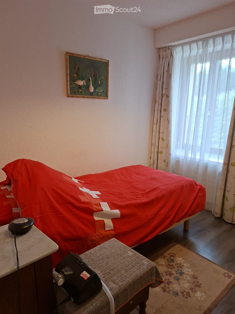 A cozy bedroom with a bed covered in a red blanket featuring white crosses, a nightstand with a black device, a framed picture on the wall, sheer curtains, and a small stool with a pet carrier on it.