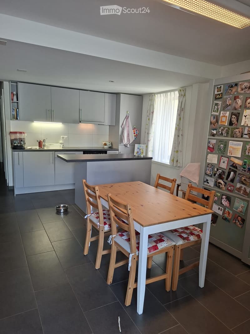 Modern kitchen with grey cabinets, a central island, a wooden dining table with four chairs, and a refrigerator adorned with photos and notes.