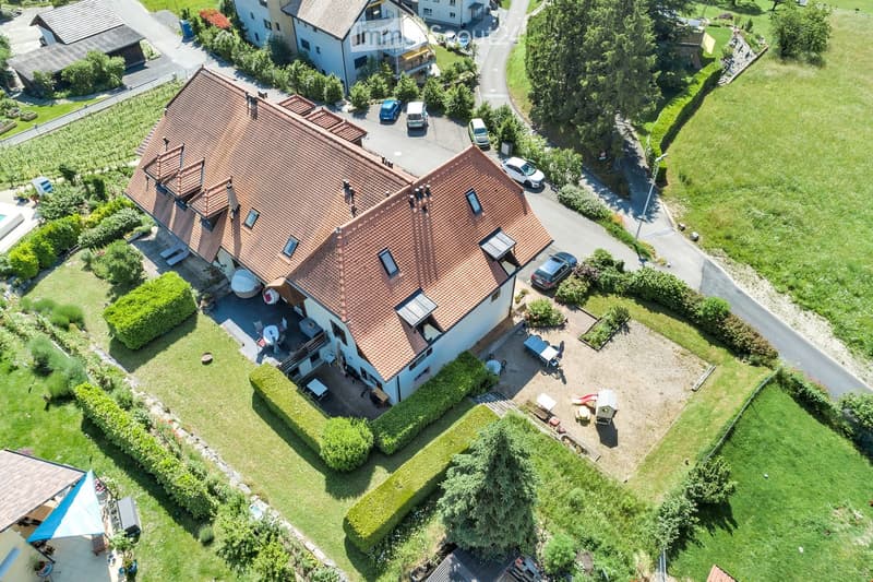 Aerial view of a two-story house with a red-tiled roof, surrounded by a well-maintained garden, driveway, and parking area. The house features a patio with outdoor furniture and a swimming pool in the yard.