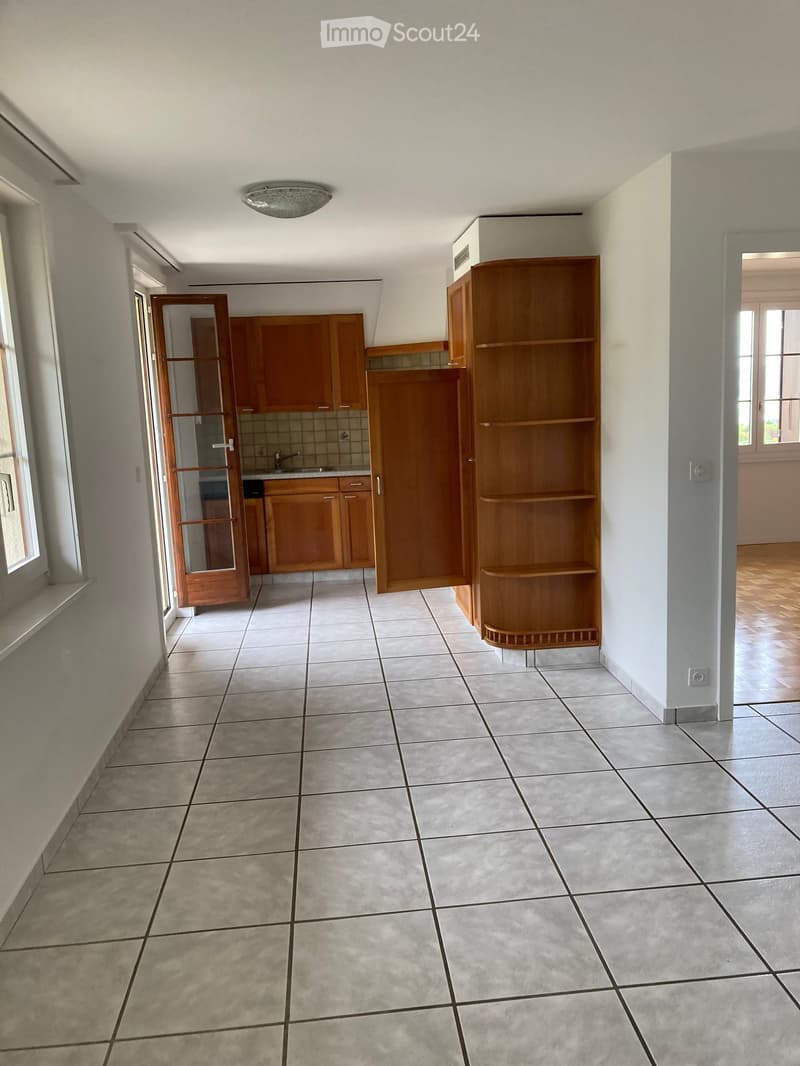 Empty room with tiled floor, wooden shelving unit, and an open kitchen area with cabinets and sink.