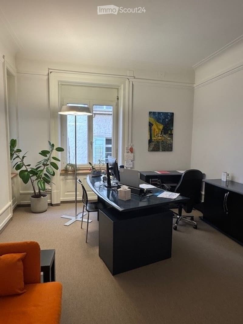 Modern office space with a large desk, laptop, office chair, sofa, potted plant, and a floor lamp. The room features large windows and a neutral color palette.