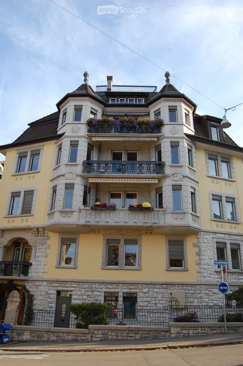 A three-story yellow villa with ornate balconies, decorative stonework, and flower boxes. The building features multiple windows, a stone base, and a roof with decorative turrets.