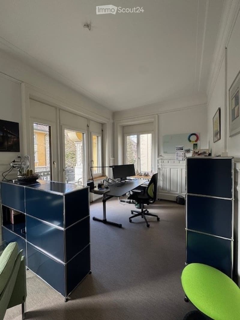 Modern office space with a large desk, ergonomic chair, computer, and storage cabinets. Large windows provide natural light, and the room features contemporary furniture.